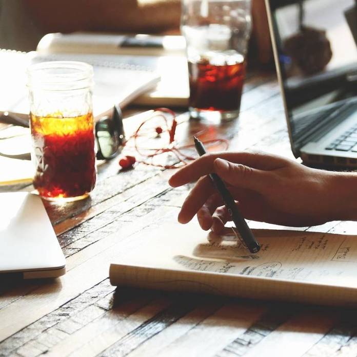 Close-up of table in a Cornish creative writing workshop led by Gail Owen, writer and creative writing teacher. On the table are a notebook and hand holding a pen, other notebooks and a laptop plus refreshments - sugary drinks for energy.