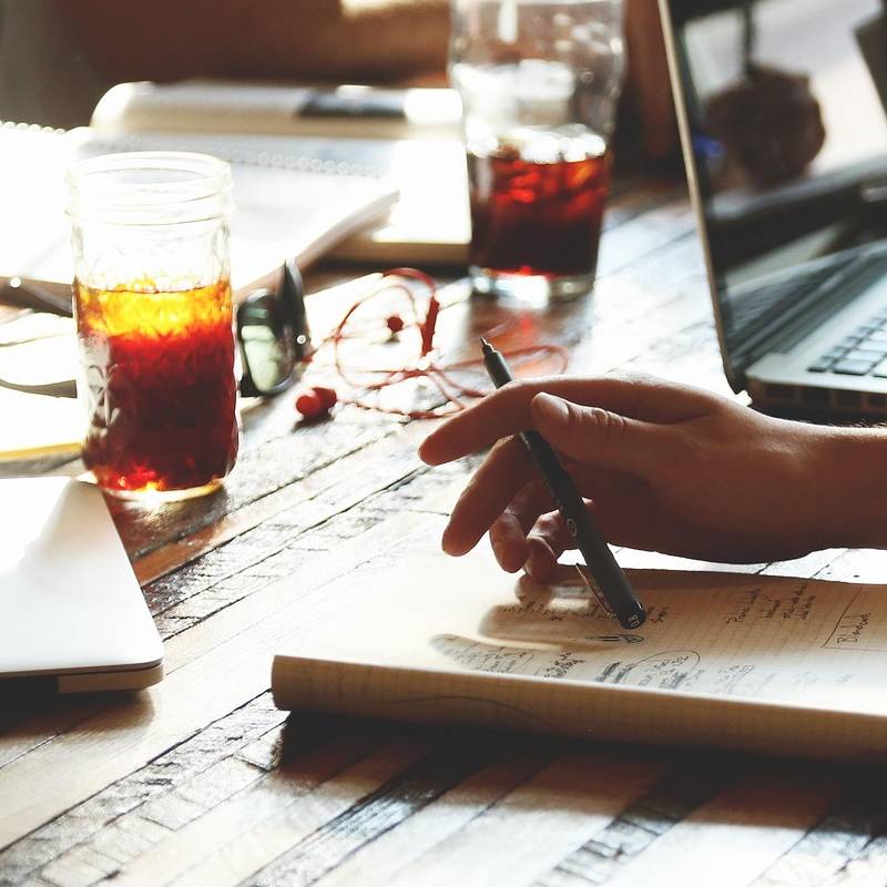 Close-up of table in a creative writing workshop: a notebook and hand holding a pen, other notebooks and a laptop plus refreshments - sugary drinks for energy.