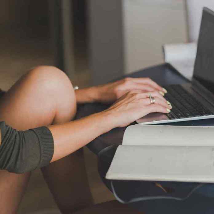 Girl typing on laptop, with one knee resting on her table, inspired by the Cornwall College creative writing course.