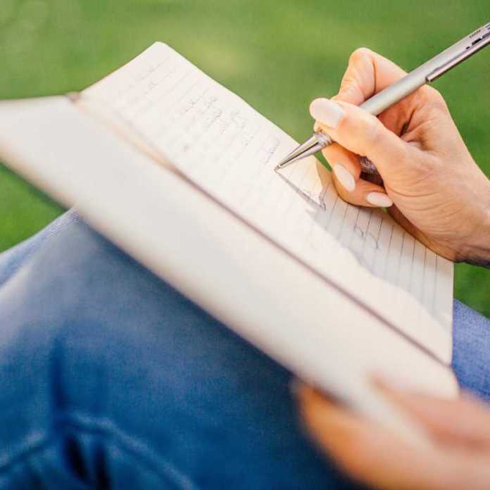 Girl, sitting on the grass, with notebook on her knees, writing her story. An idyllic picture of a Cornish creative writing class.
